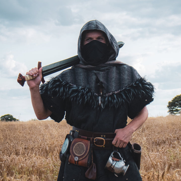 Person in medieval attire holding a sword in a field with a cloudy sky.