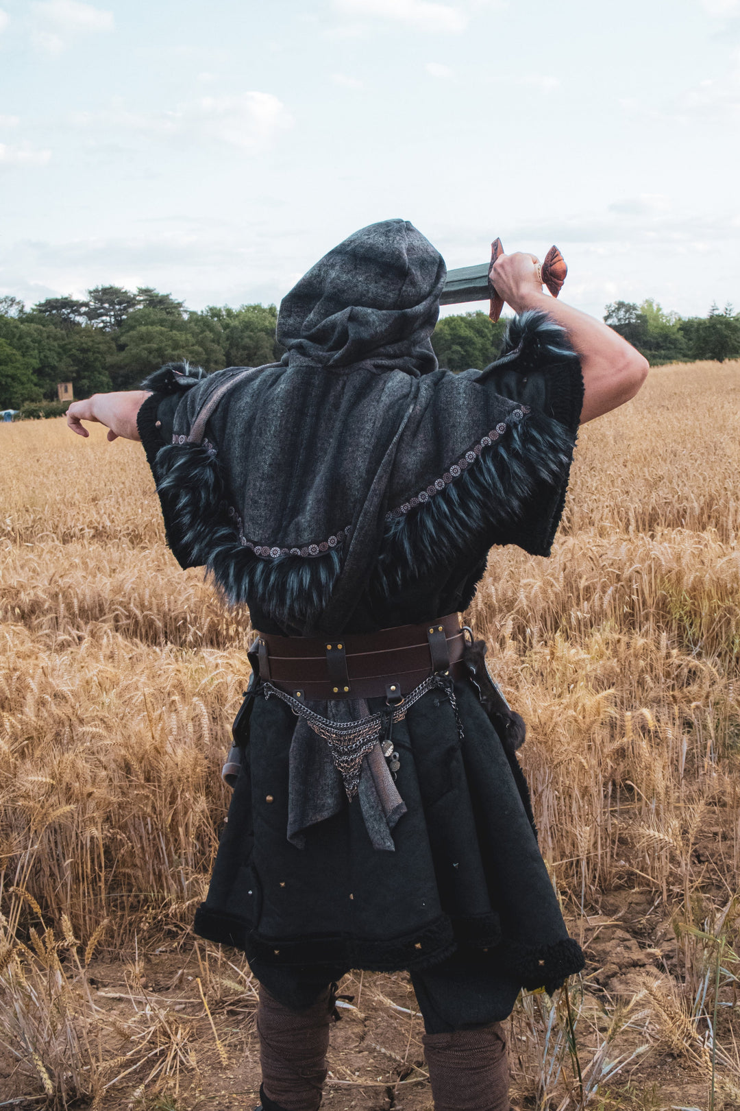 Person in a hooded cloak holding a sword in a field with a cloudy sky.