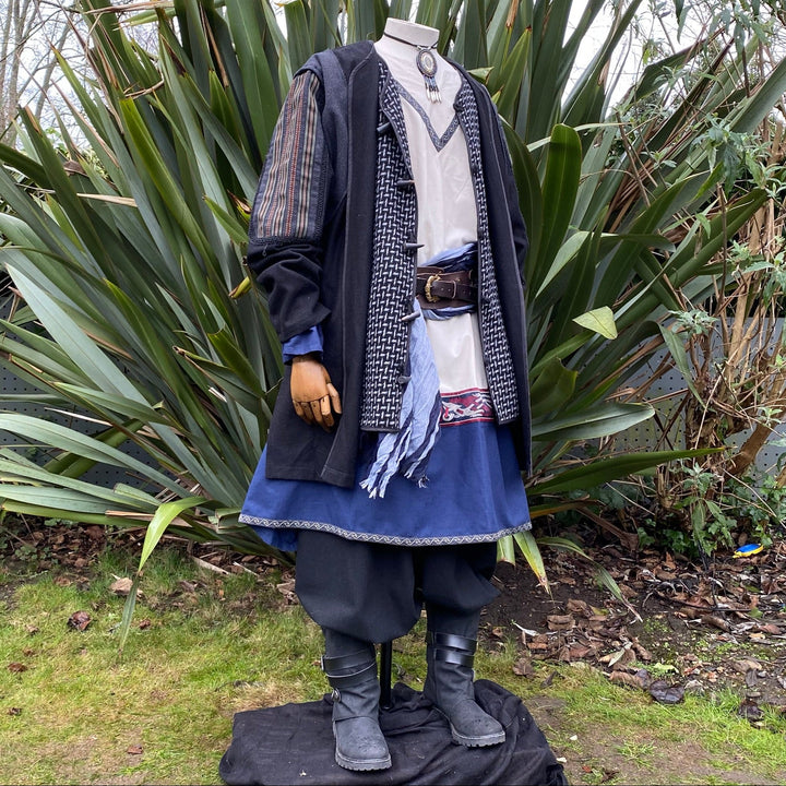 Medieval LARP costume side view, featuring a black and grey wool jacket with ornate panels, a wool hood, tunic, pants, sash and necklace. For Renaissance Faire and historical roleplay events.
