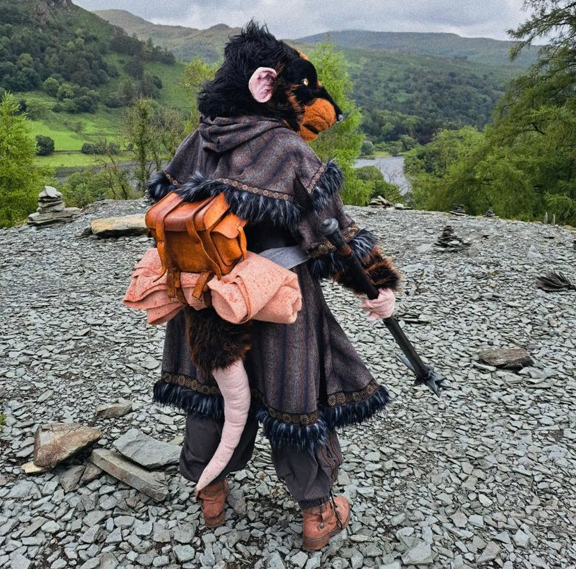 Person in a costume resembling a rat with backpack and walking stick, standing on a rocky landscape with green hills and water in the background.