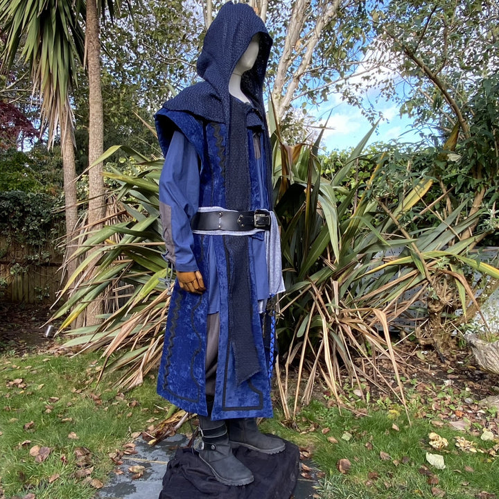 Medieval LARP costume side view, featuring a blue suede-effect panel waistcoat, wool hood, two-tone cotton tunic and wool sash. For Renaissance Faire and historical roleplay events.