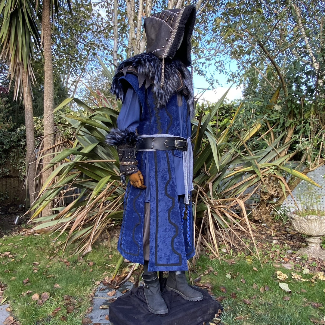 Medieval LARP costume side view, featuring a blue suede-effect panel waistcoat, black faux-leather hood and vambraces, tunic, leather belt and sash. For Renaissance Faire and historical roleplay events.