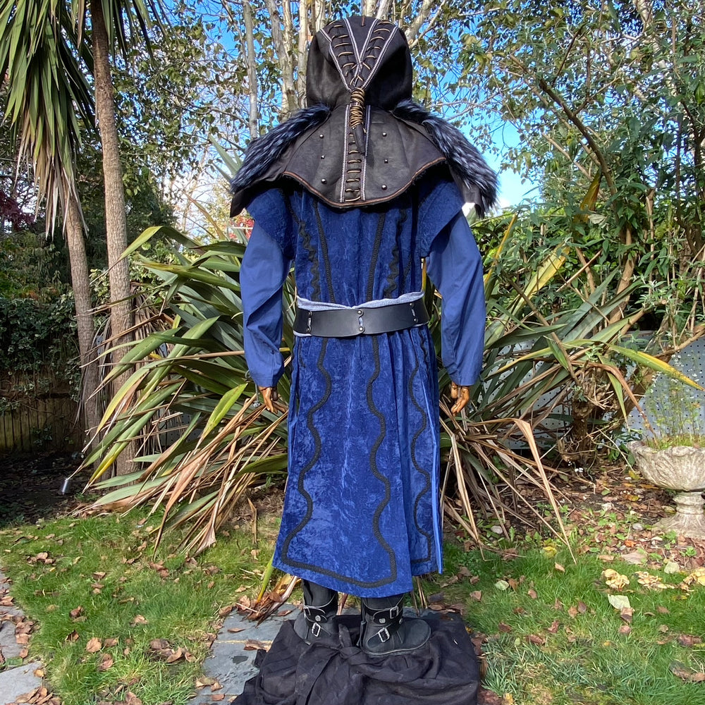 Medieval LARP costume back view, featuring a blue suede-effect panel waistcoat, black faux-leather hood, two-tone cotton tunic and wool sash. For Renaissance Faire and historical roleplay events.