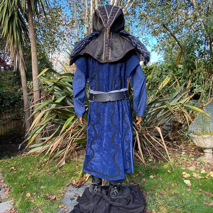 Medieval LARP costume back view, featuring a blue suede-effect panel waistcoat, black faux-leather hood, two-tone cotton tunic and wool sash. For Renaissance Faire and historical roleplay events.