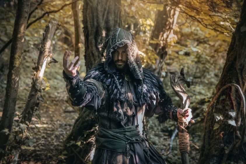 Dark medieval LARP outfit with hood, vambraces, and necklace in forest setting.