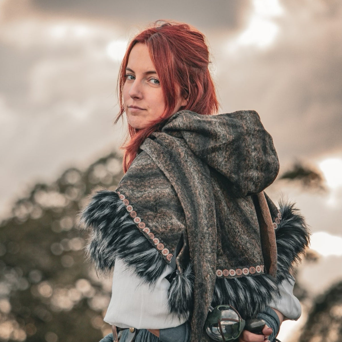 Red-haired figure in side profile with medieval mohair hood, grey sky background.