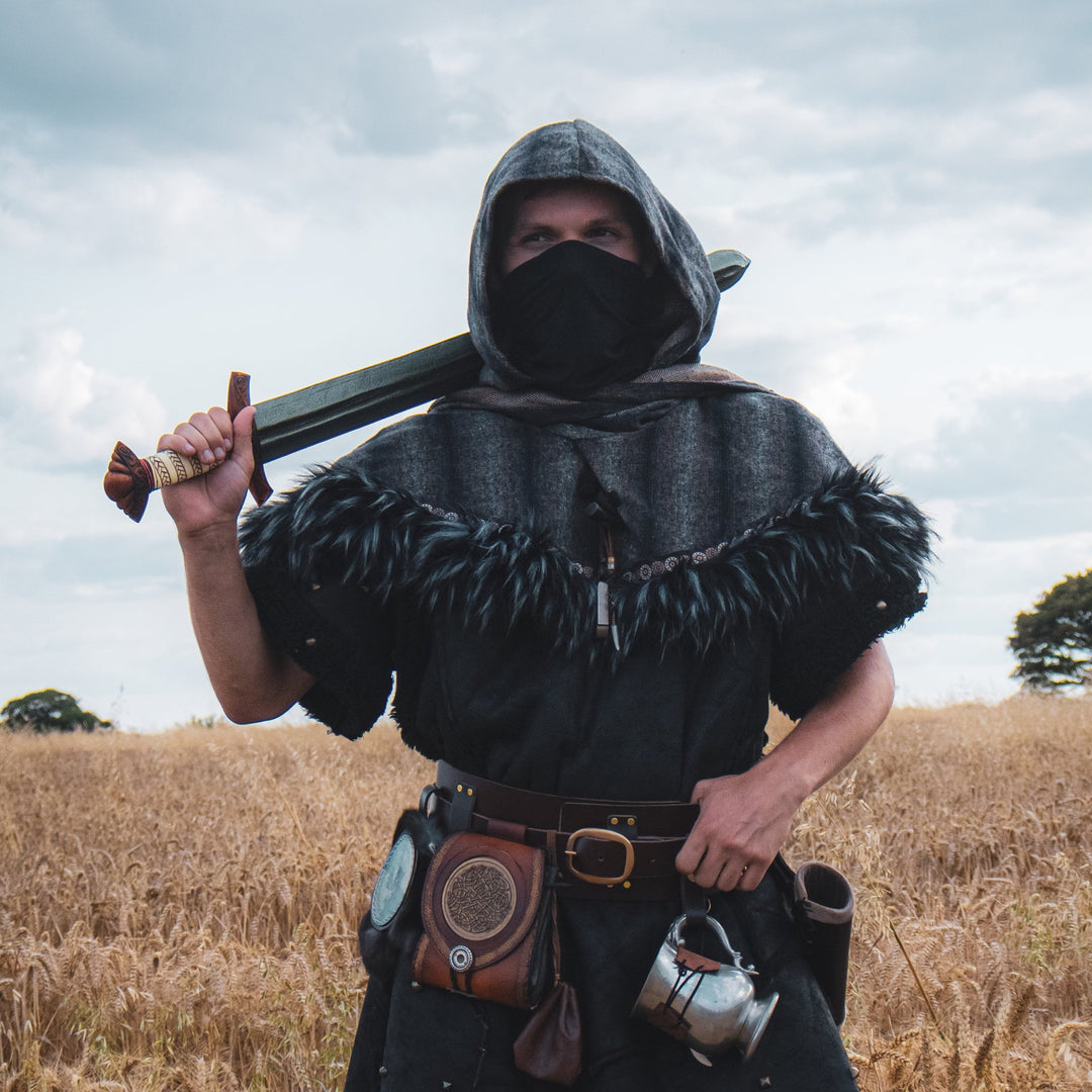 Person in medieval attire holding a sword in a field with a cloudy sky.