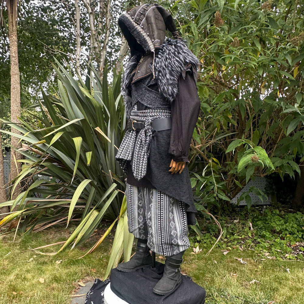 Medieval LARP costume side view, includes faux leather hood, black and white waistcoat with asymmetrical front panel and matching sash, wool hood, shirt, trousers and leather belt for roleplay events.