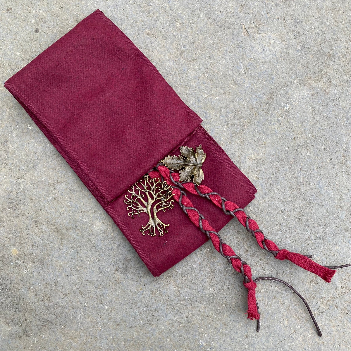Braided crimson LARP sash with brass leaf and tree accents on stone backdrop.