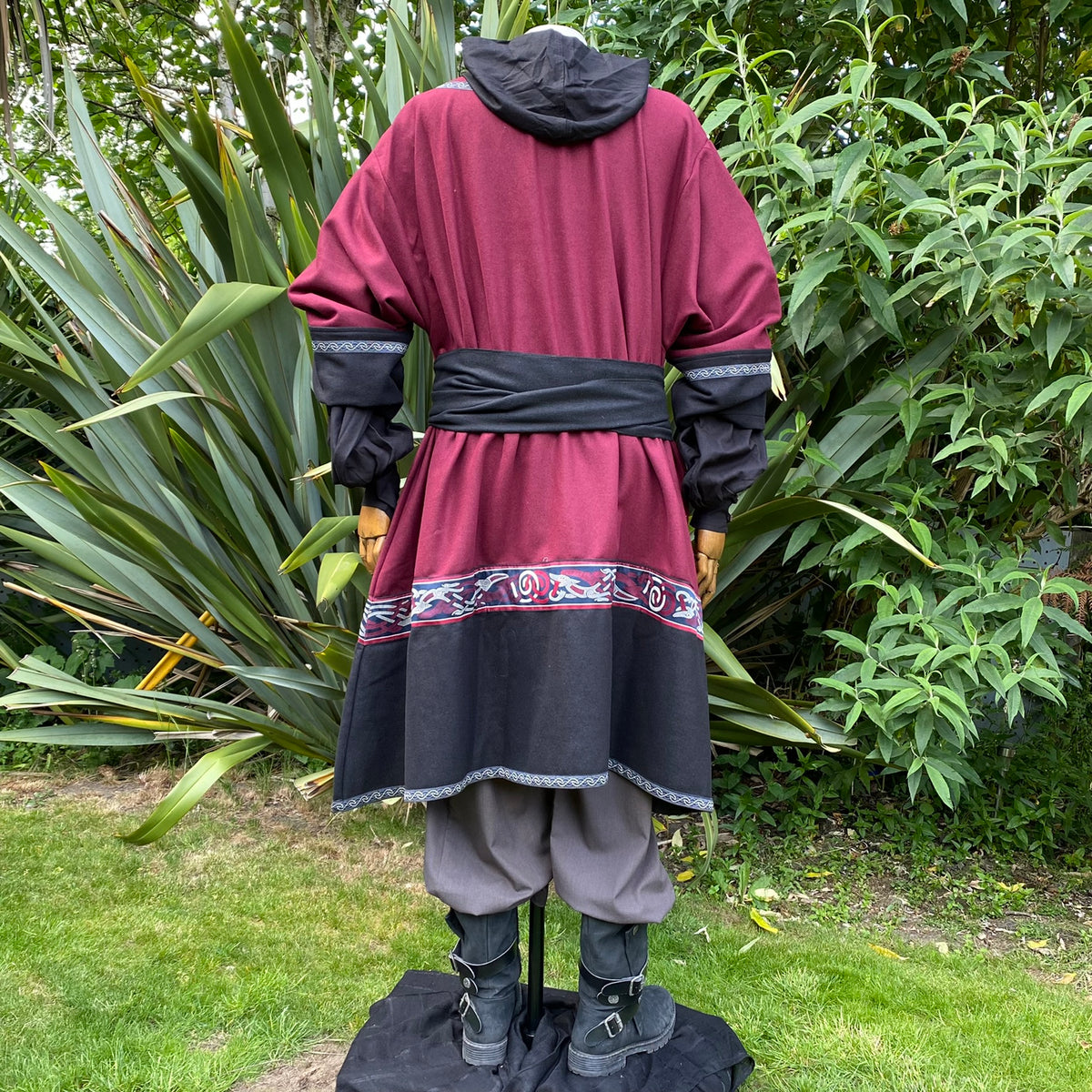 Back view of a red-black medieval tunic with hood, sash, and decorative trim.