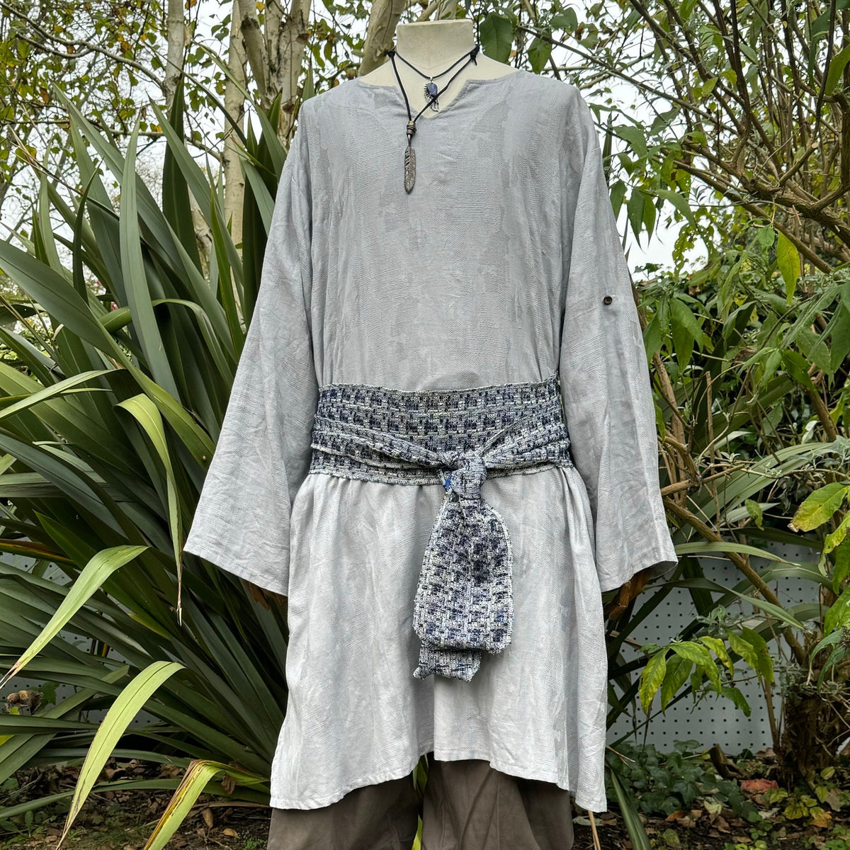 Front view of grey LARP tunic with patterned sash among garden greenery.