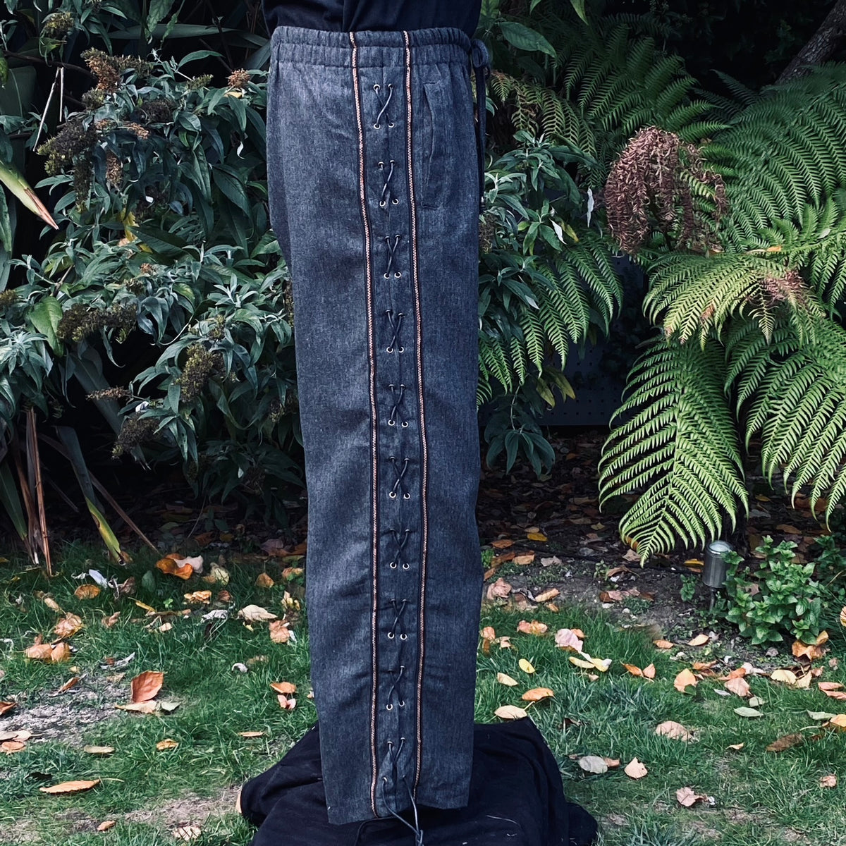 Side view of grey wool pants with wide decorative trim and cross-lace detailing no boots.