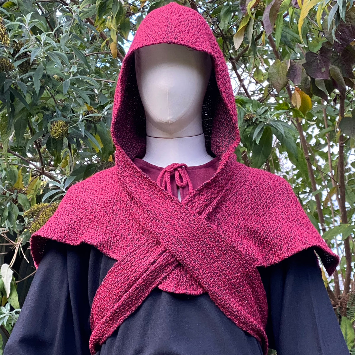LARP medieval outfit with red hood and crisscross sash against foliage backdrop.