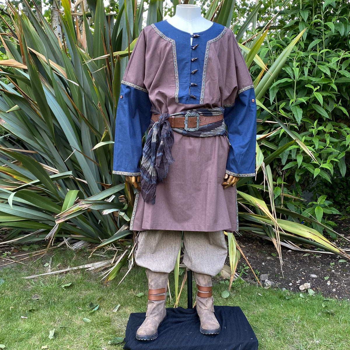 Medieval LARP tunic in garden, brown-blue, belted, front view with boots.