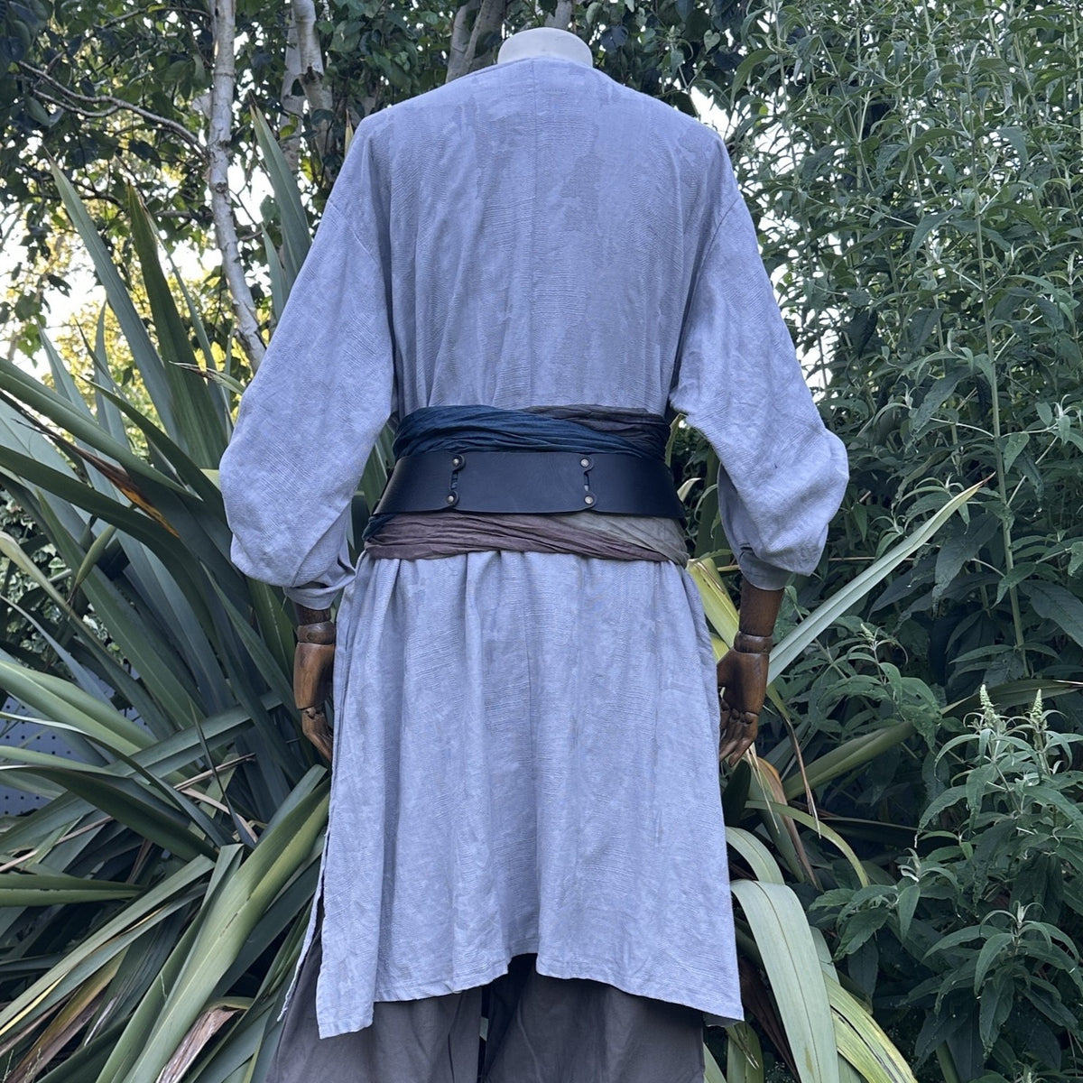 Viking LARP tunic set, back view, grey fabric, leather sash, lush foliage background.