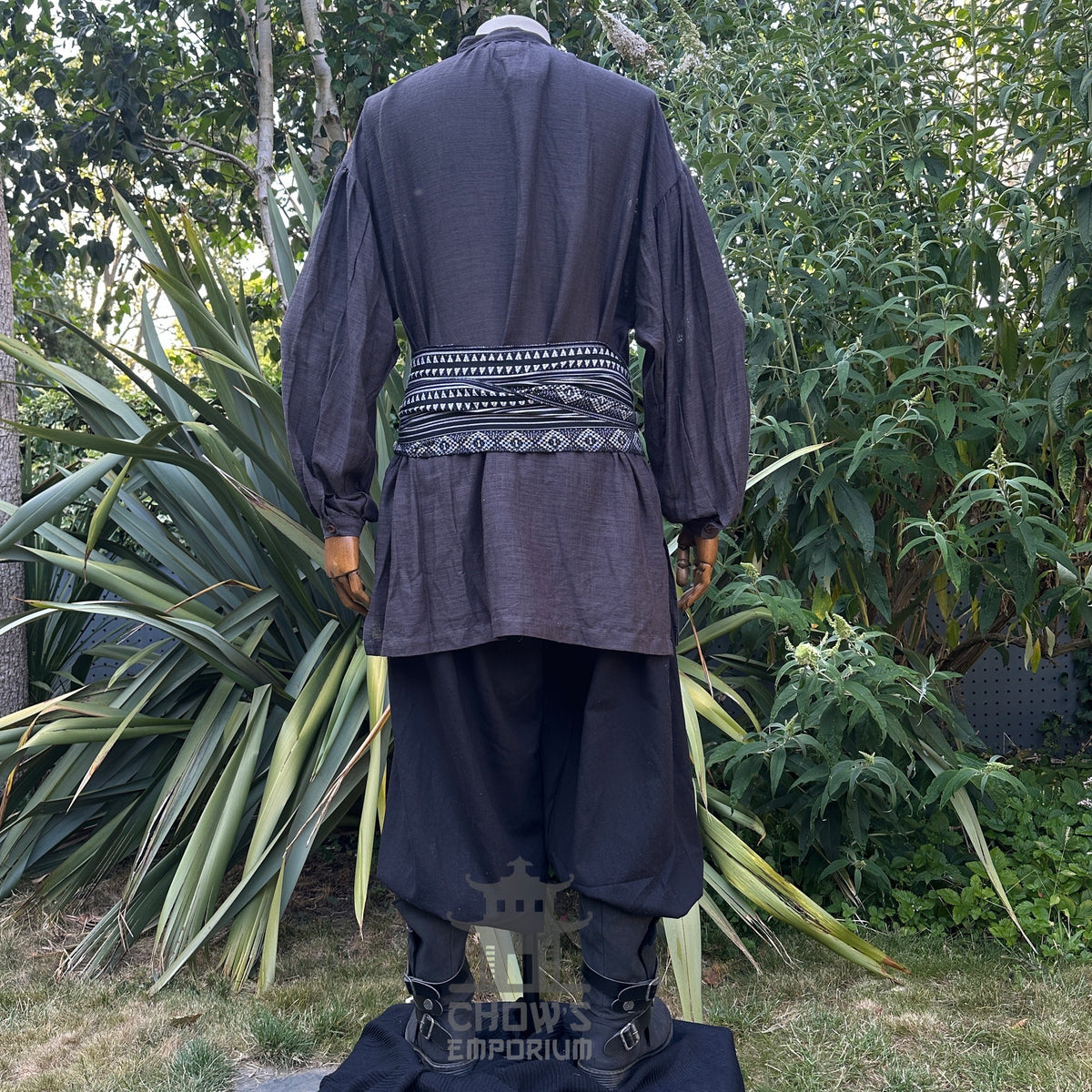 Back view of a medieval LARP set, black waistcoat, grey shirt, patterned sash.