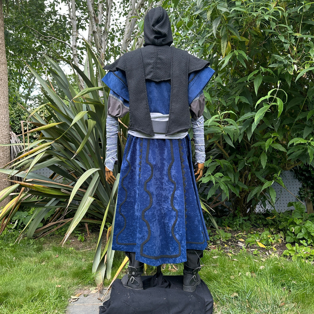 Medieval LARP costume back view, featuring a blue suede-effect panel waistcoat, wool hood, two-tone cotton tunic, arm wraps and sash. For Renaissance Faire and historical roleplay events.