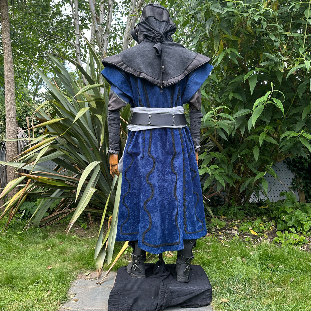 Medieval LARP costume back view, featuring a blue suede-effect panel waistcoat, black layered hood, tunic, trousers, arm wraps, leather belt and sash. For Ren Faire and historical roleplay events.