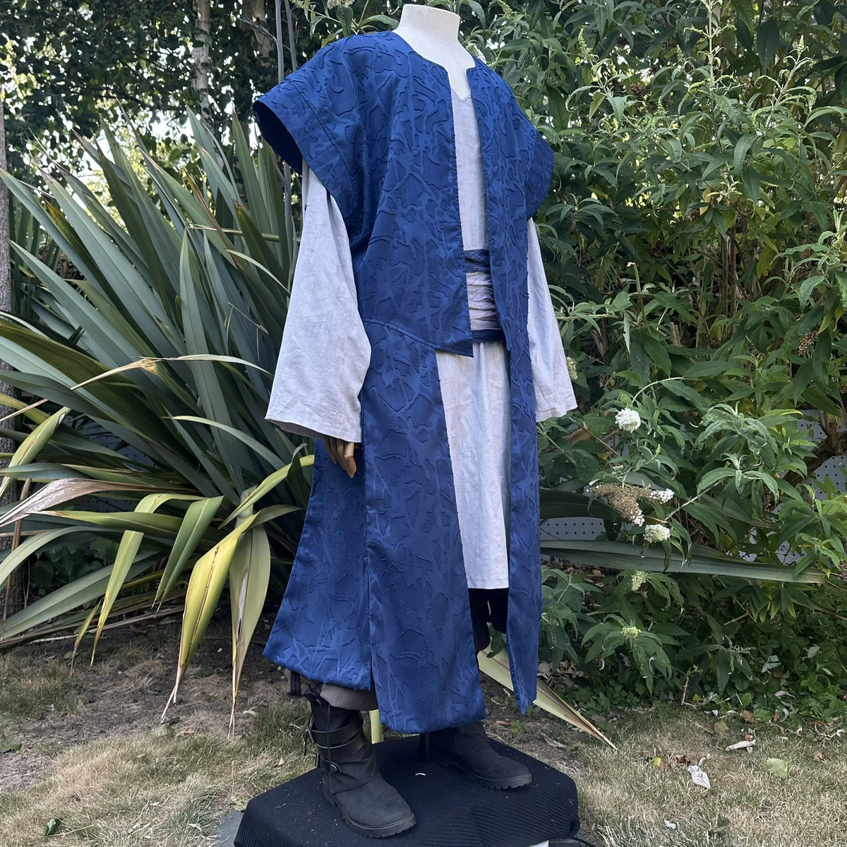 Blue panelled waistcoat worn open over a shirt with a sash at the waist, hood down, three-quarter view. Shirt and sash sold separately.