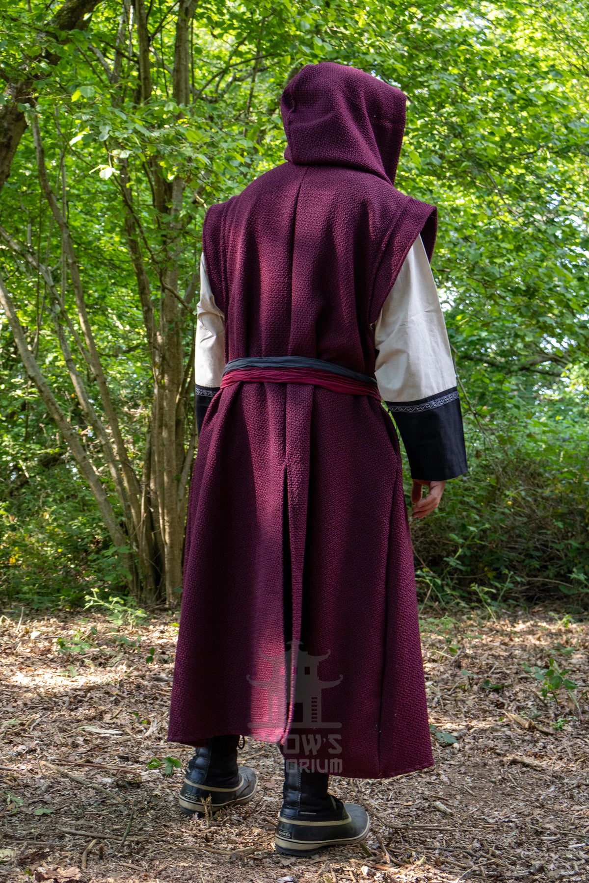 A person viewed from behind wearing a medieval-style hooded robe in a wooded outdoor setting. The robe is deep burgundy with a dark waist sash and contrasting white and black sleeves, paired with dark boots.