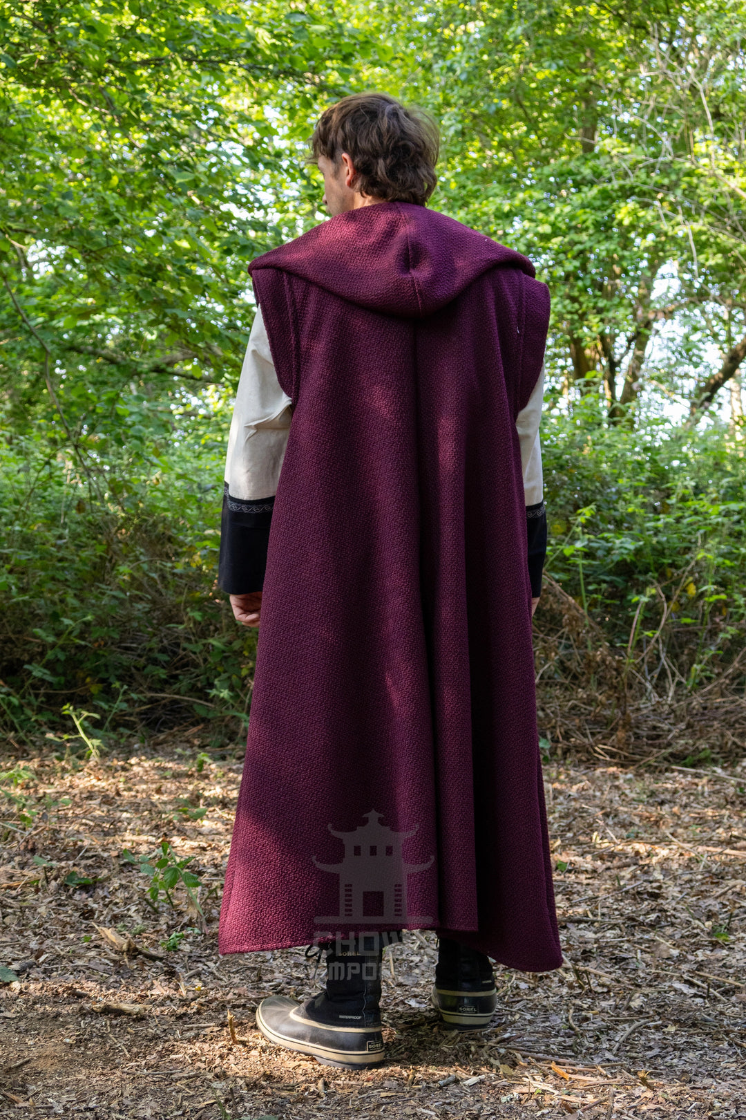 A person standing outdoors in a wooded area, viewed from behind, wearing a long burgundy hooded cloak over a white and black outfit. The background consists of green trees and brown forest floor.