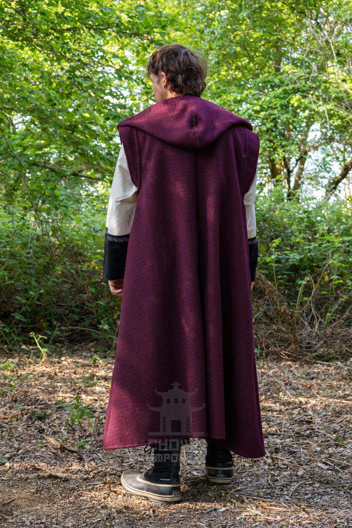 A person standing outdoors in a wooded area, viewed from behind, wearing a long burgundy hooded cloak over a white and black outfit. The background consists of green trees and brown forest floor.