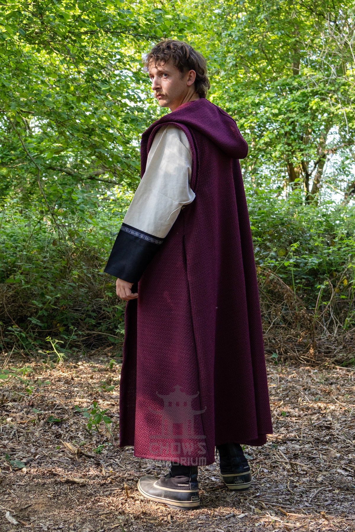 A man wearing a long burgundy medieval-style hooded cloak over a white and black sleeveless tunic, standing in a wooded outdoor setting.