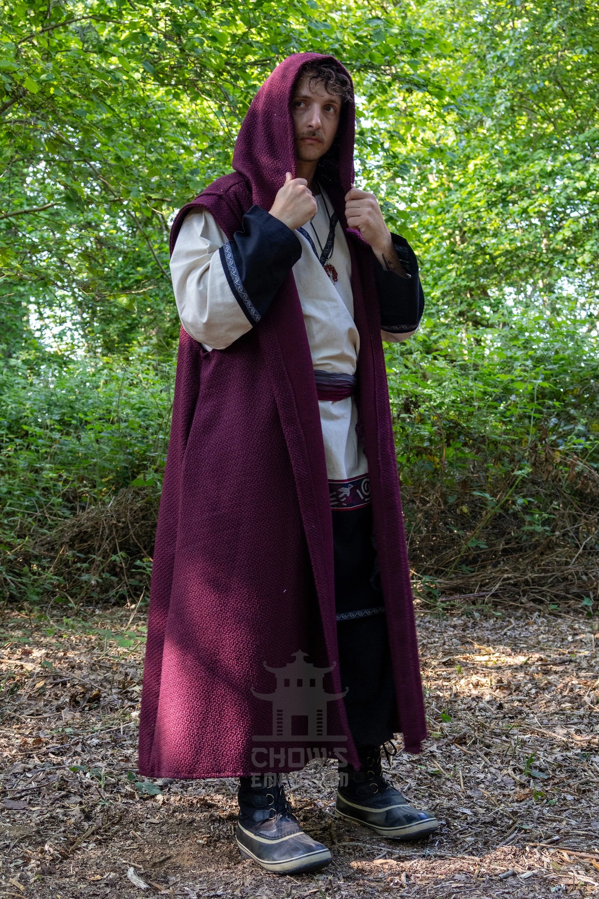 A man wearing a medieval-style costume stands outdoors in a wooded setting. The outfit includes a long burgundy hooded cloak over a cream tunic with black sleeves, black trousers, and dark boots.