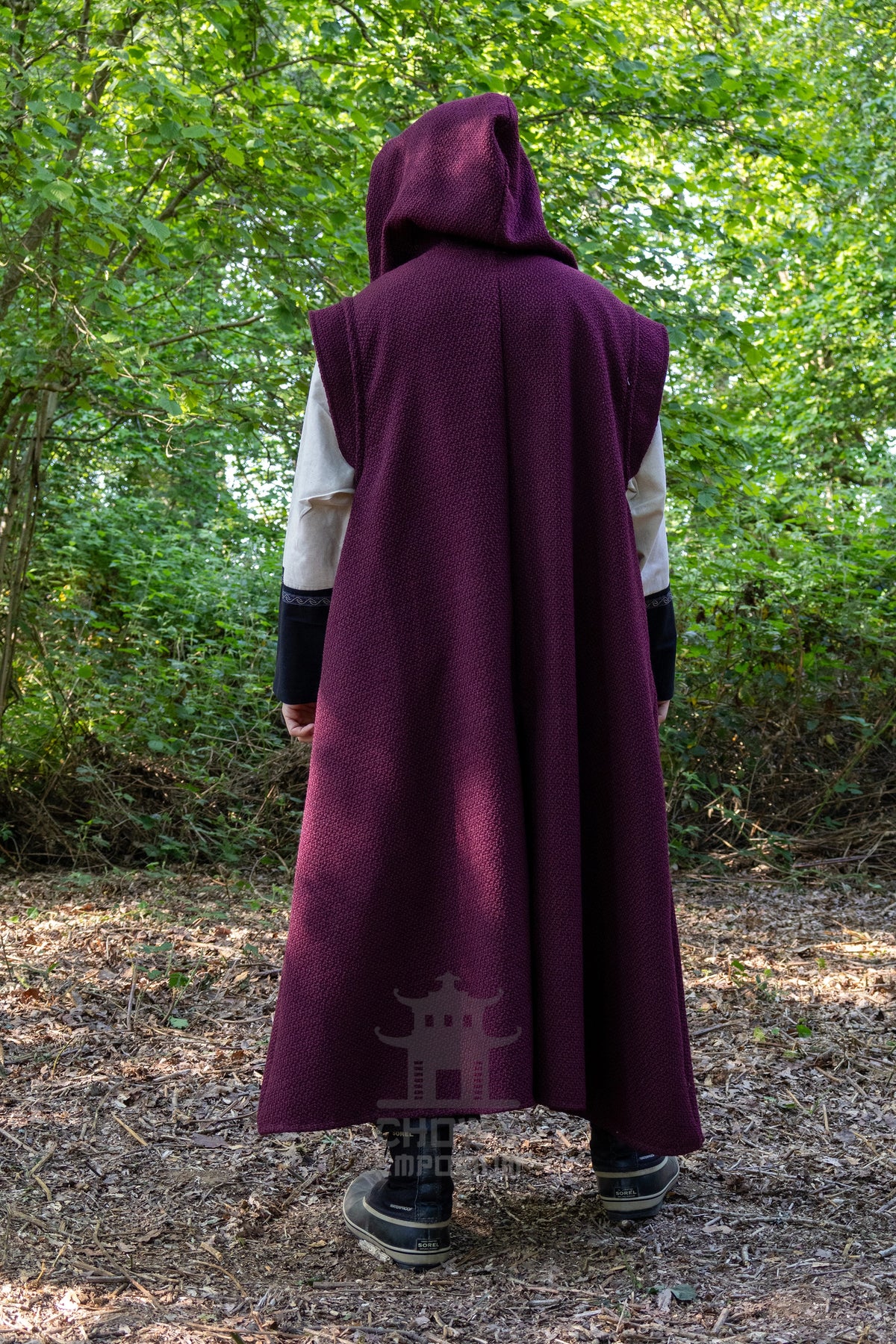 A person viewed from behind wearing a long, sleeveless hooded robe in deep burgundy purple over a white shirt and black pants, standing in a wooded outdoor setting.