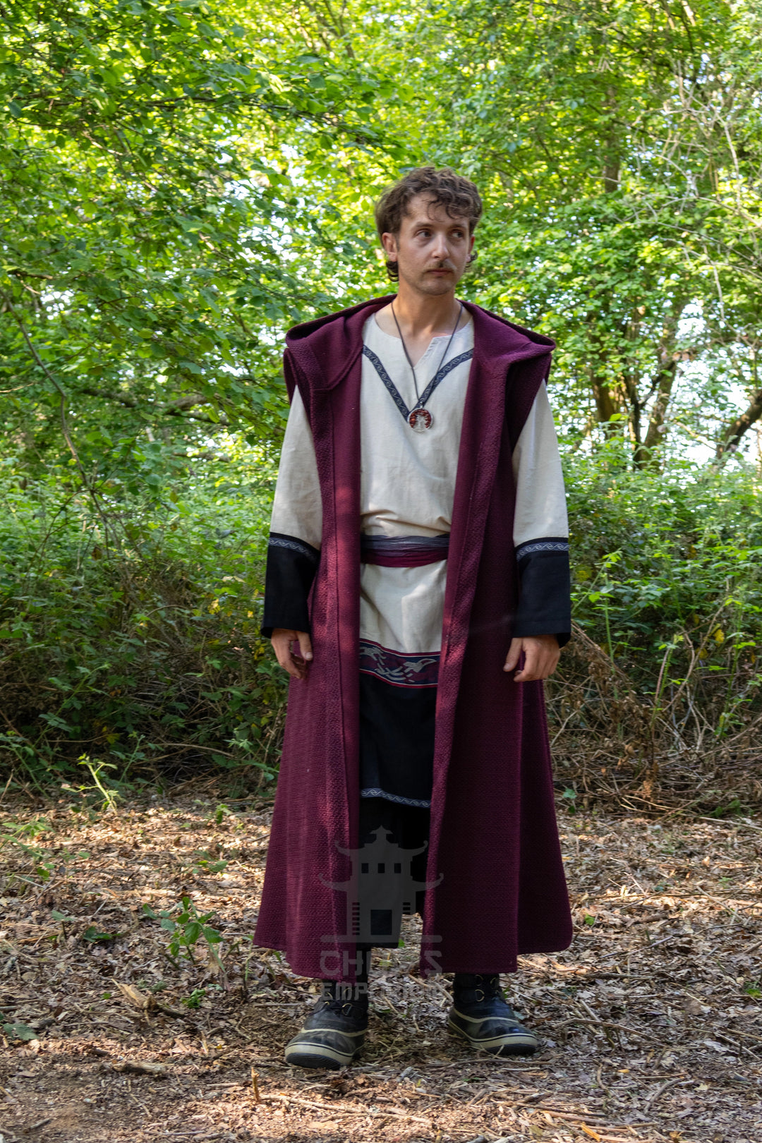 A man wearing a Viking-style costume stands in a wooded outdoor setting. The costume features a long burgundy outer robe over a cream tunic with black sleeve accents, a dark skirt, and a necklace.