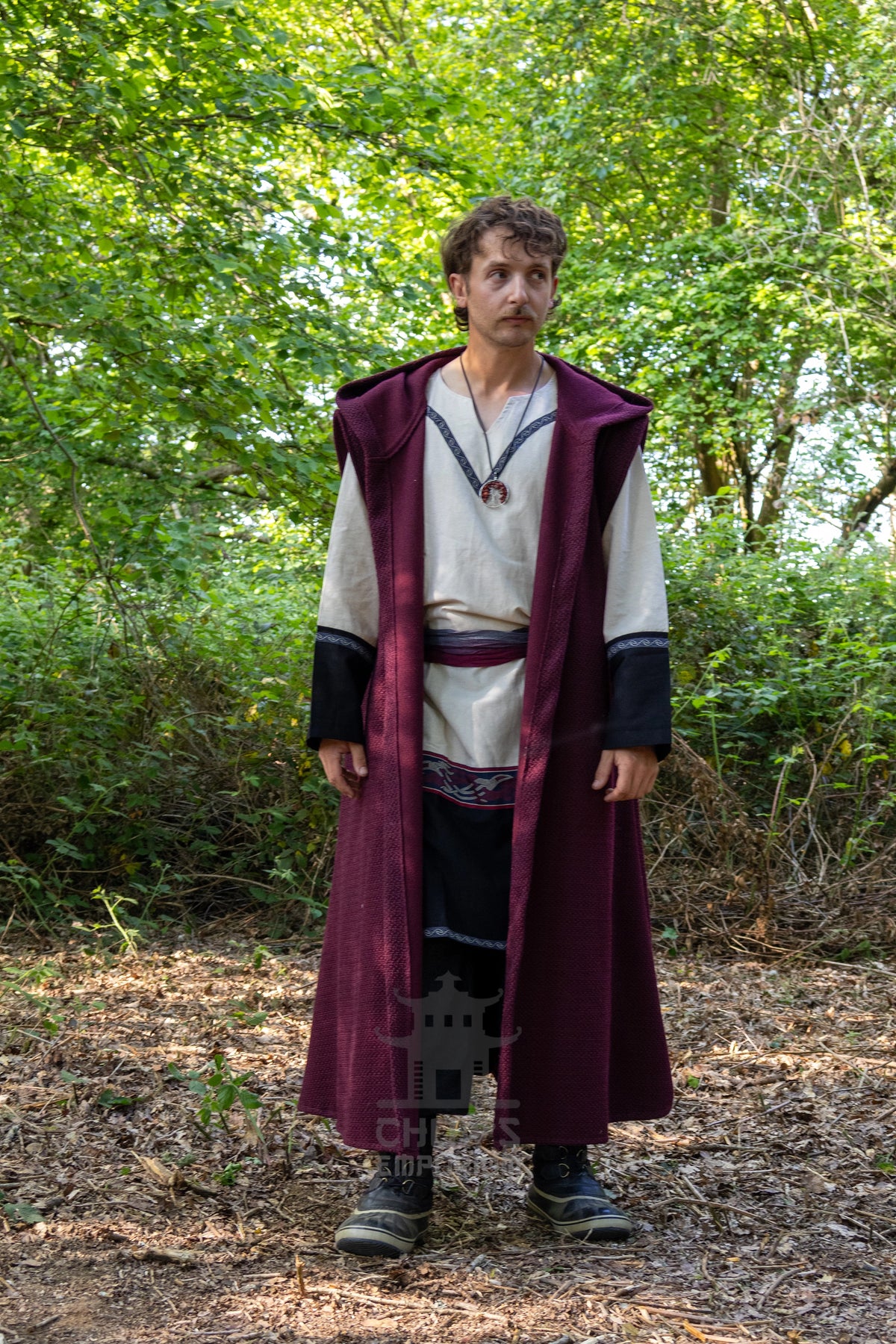 A man wearing a Viking-style costume stands in a wooded outdoor setting. The costume features a long burgundy outer robe over a cream tunic with black sleeve accents, a dark skirt, and a necklace.