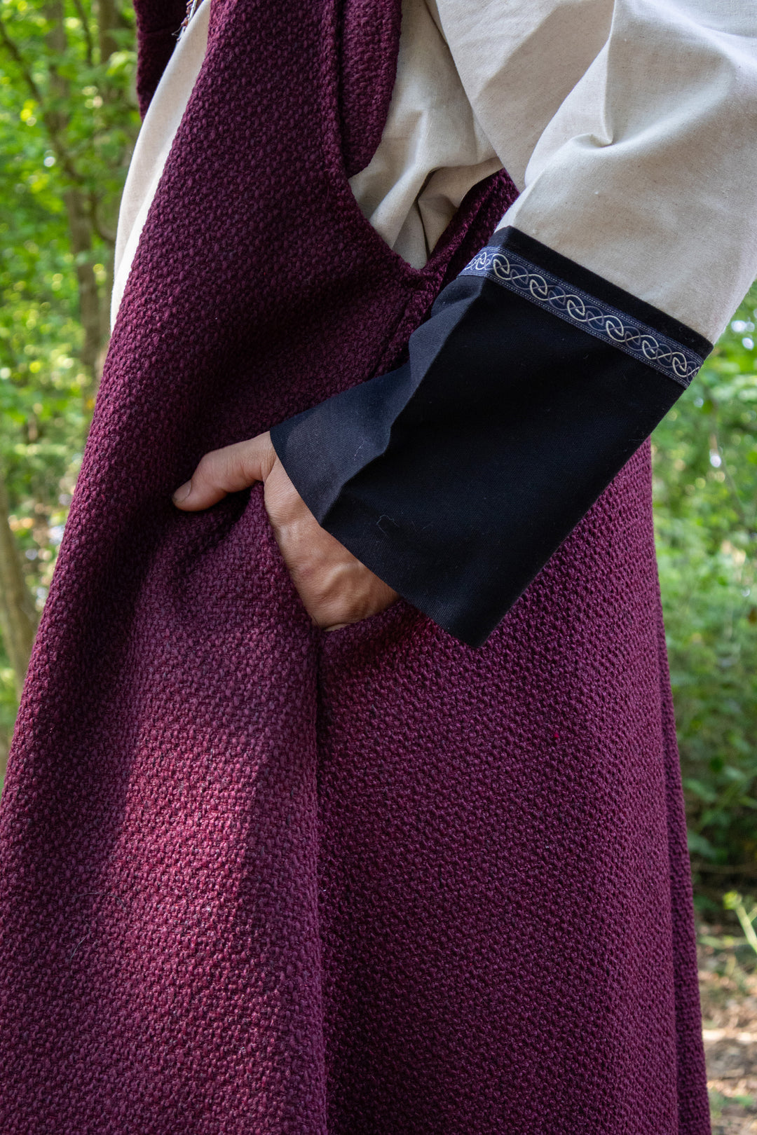 A close-up of a person wearing a textured plum purple robe or cloak over cream and black clothing, with a hand in a pocket, set against a blurred green outdoor background.