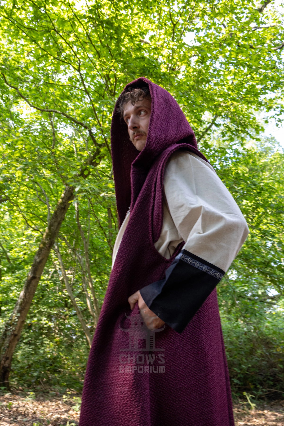 A man wearing a medieval-style long burgundy hooded cloak over a cream tunic and black sleeve, standing in a lush green forest. A watermark for 'Chow's Emporium' is visible near the bottom.