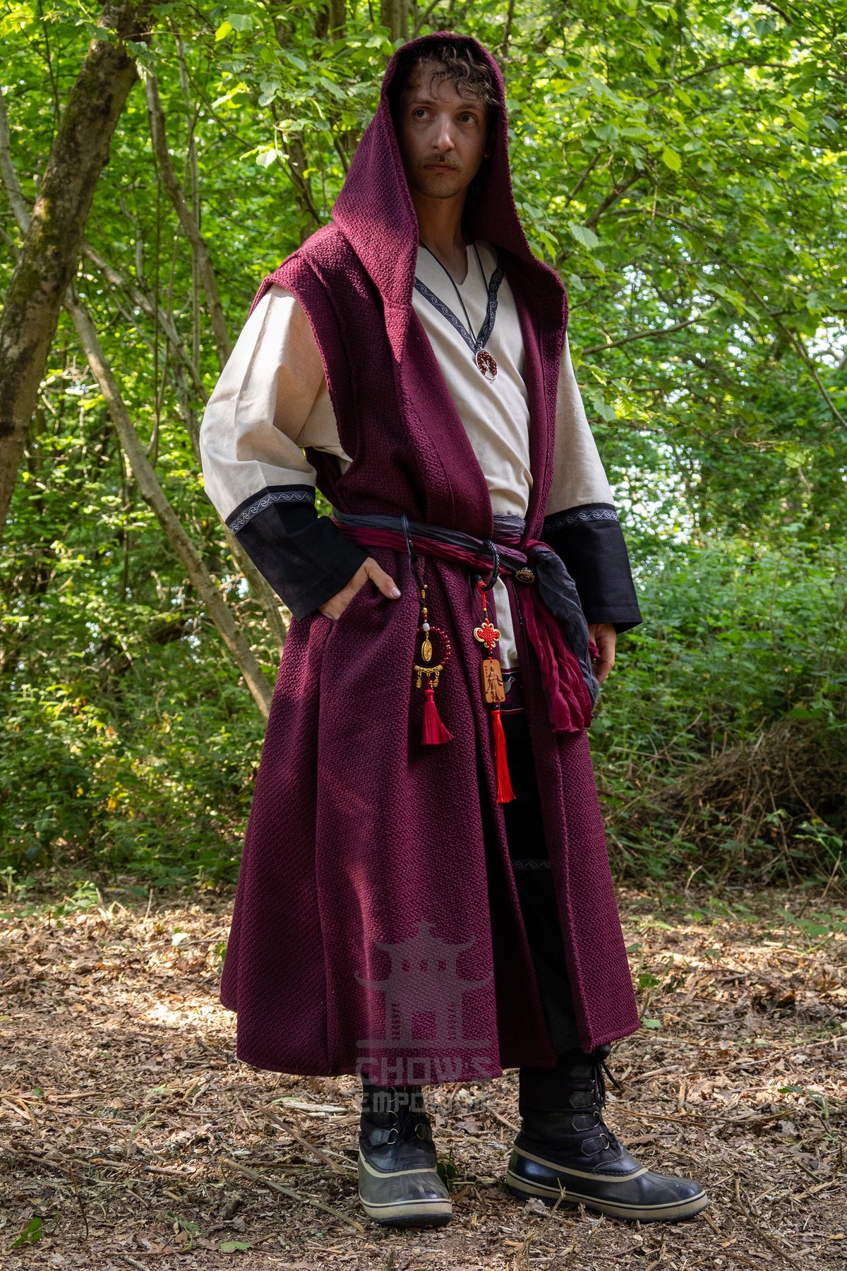 A person standing outdoors in a wooded setting wearing a long medieval-style costume with a burgundy hooded tunic over a cream shirt, dark sleeves, a dark belt with tassels, and black boots.