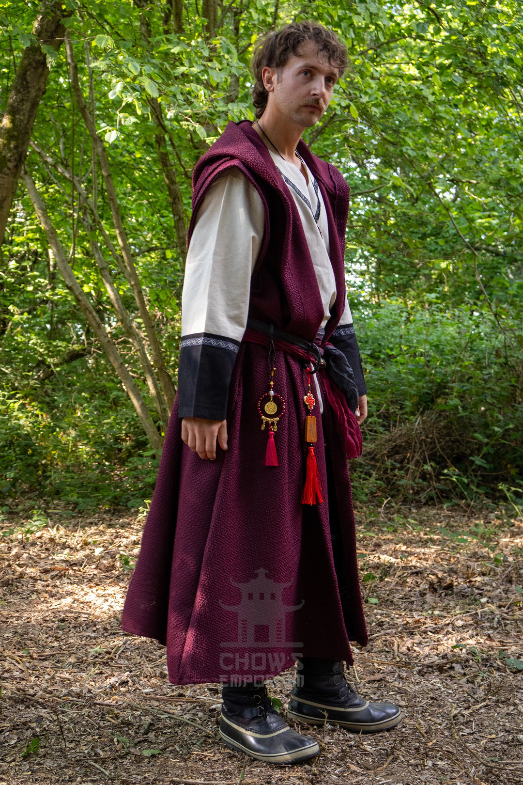 A man wearing a traditional Chinese Hanfu outfit with a deep burgundy robe, white under-sleeve, and black sash, standing in a wooded outdoor setting.
