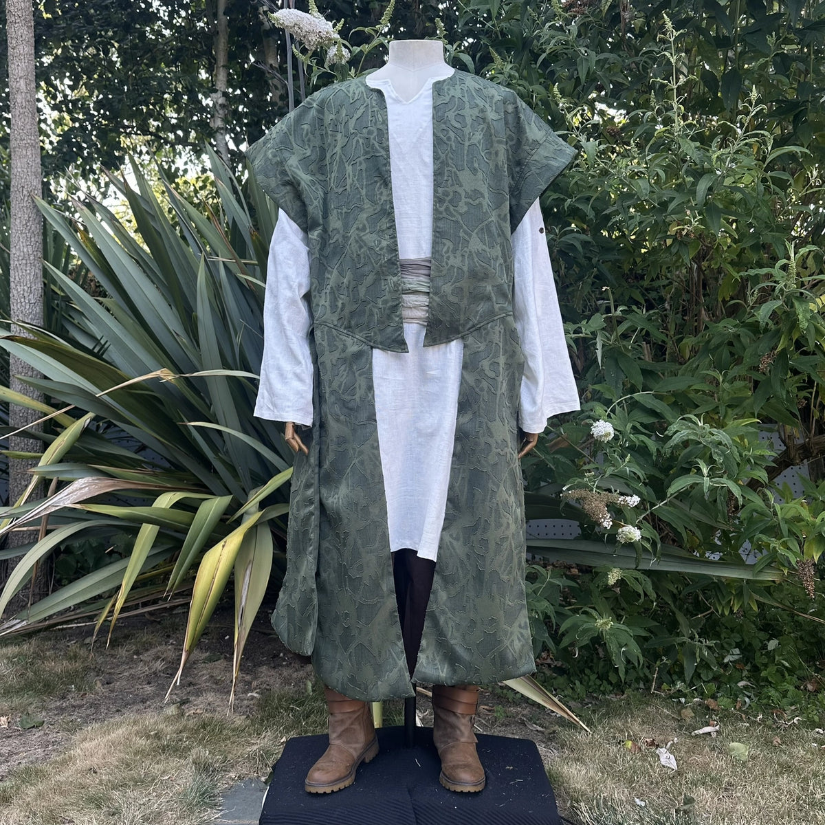 Green panelled waistcoat worn open over a shirt with a sash at the waist, hood down, three-quarter view. Shirt and sash sold separately.