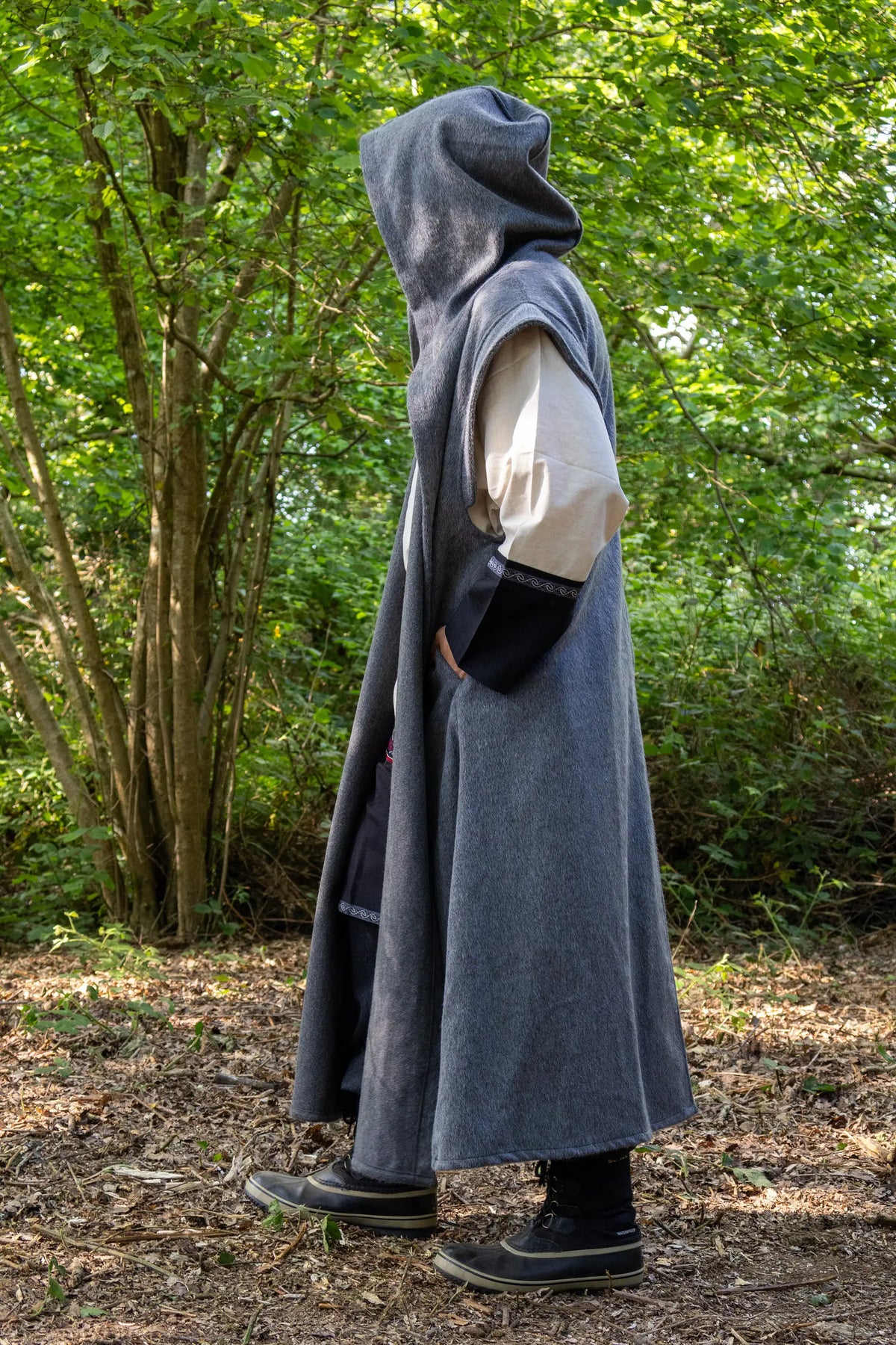 A person standing in a wooded outdoor setting wearing a long, hooded gray robe or cloak over a cream shirt and black sleeves, with black shoes and boots.