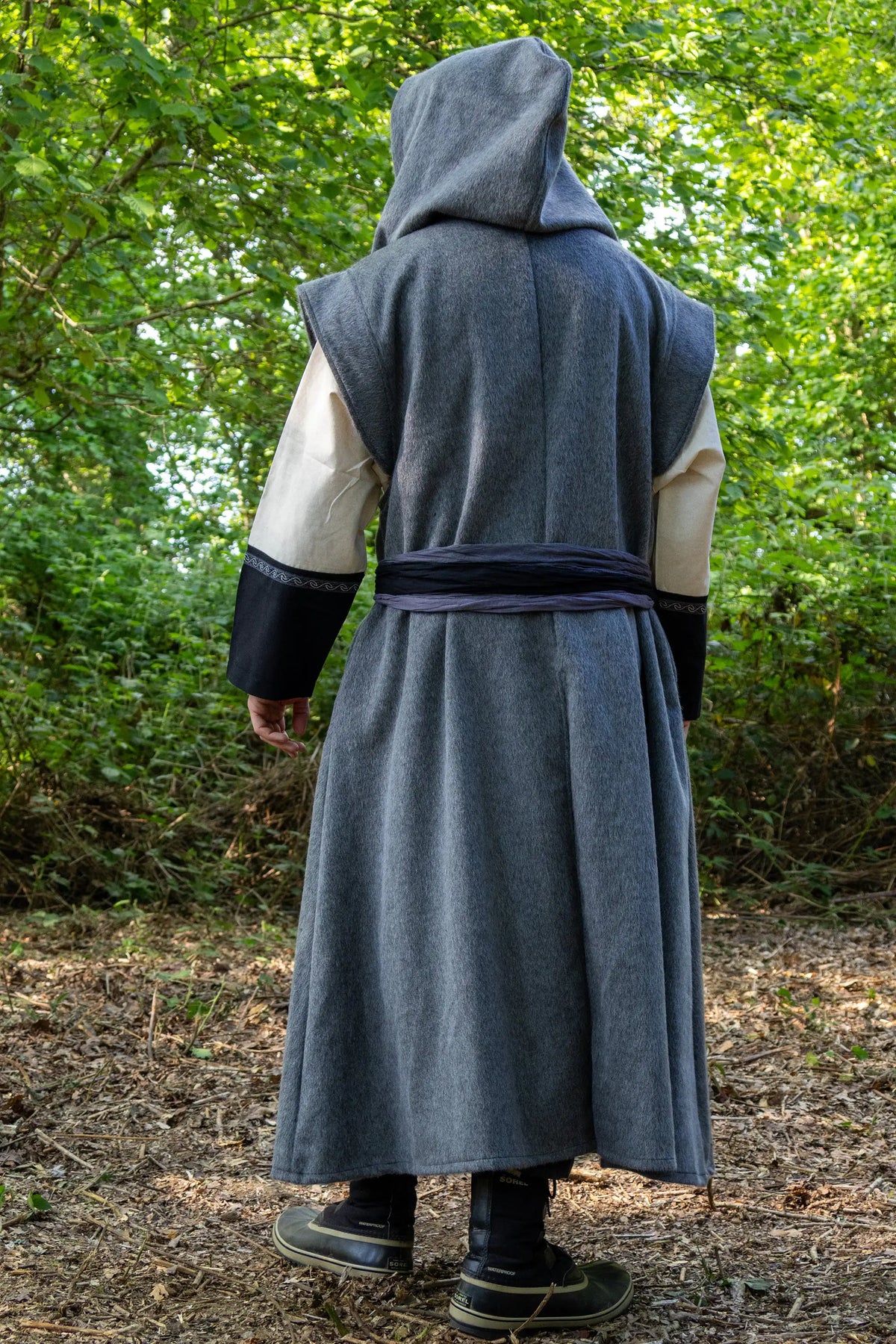 A person standing outdoors in a wooded area, viewed from the back, wearing a long hooded robe or costume in dark gray with cream sleeves and black cuffs.