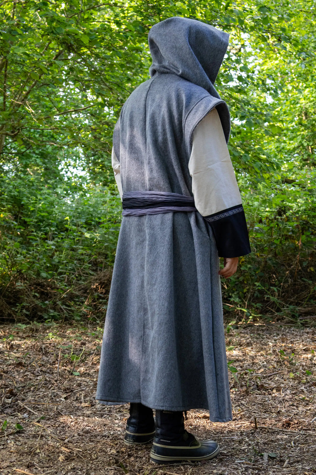 A person wearing a long gray hooded robe with a waist tie, cream sleeves, and dark cuffs, standing outdoors in a wooded area with trees and leaf-covered ground.