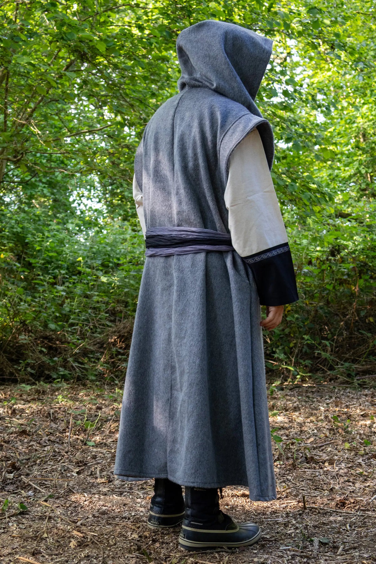 A person wearing a long gray hooded robe with a waist tie, cream sleeves, and dark cuffs, standing outdoors in a wooded area with trees and leaf-covered ground.