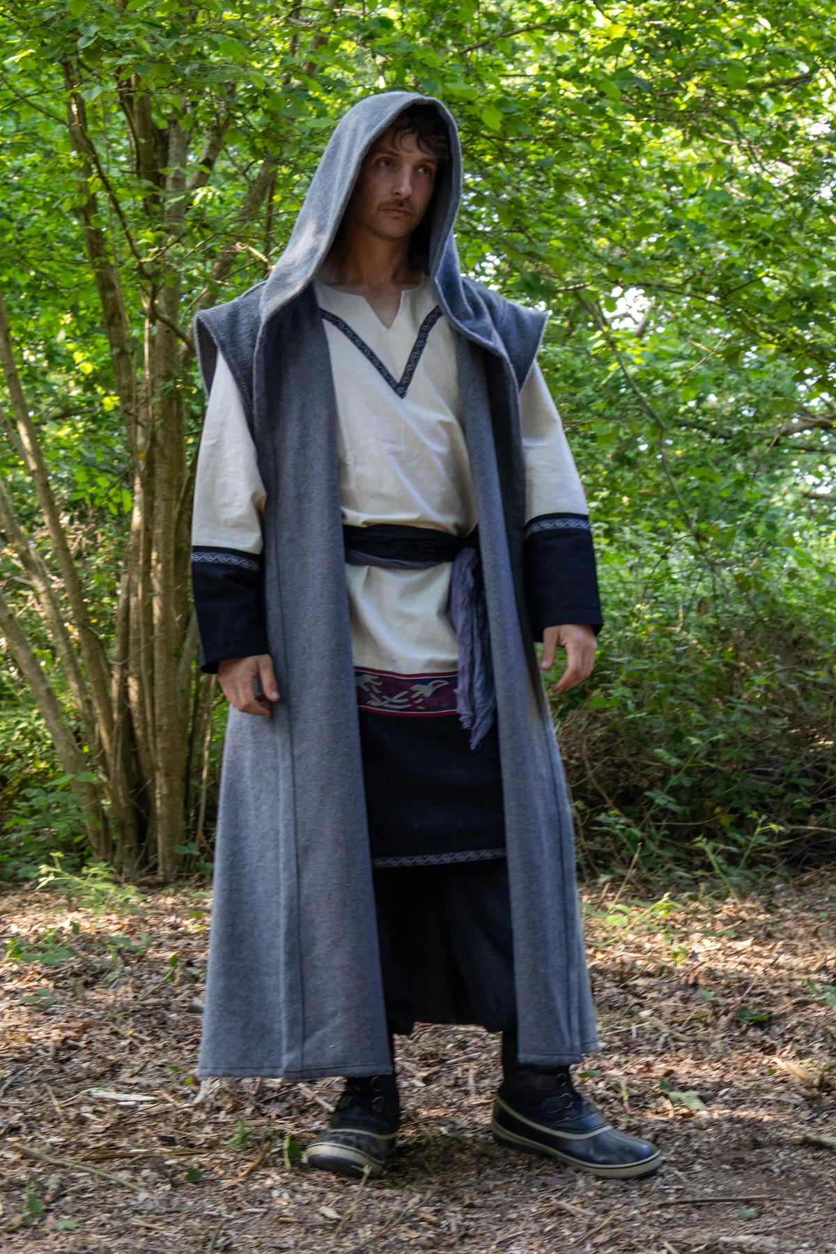 A person wearing a medieval-style long gray hooded robe over a cream tunic and dark skirt, standing outdoors in a wooded area with trees and leaf-covered ground.
