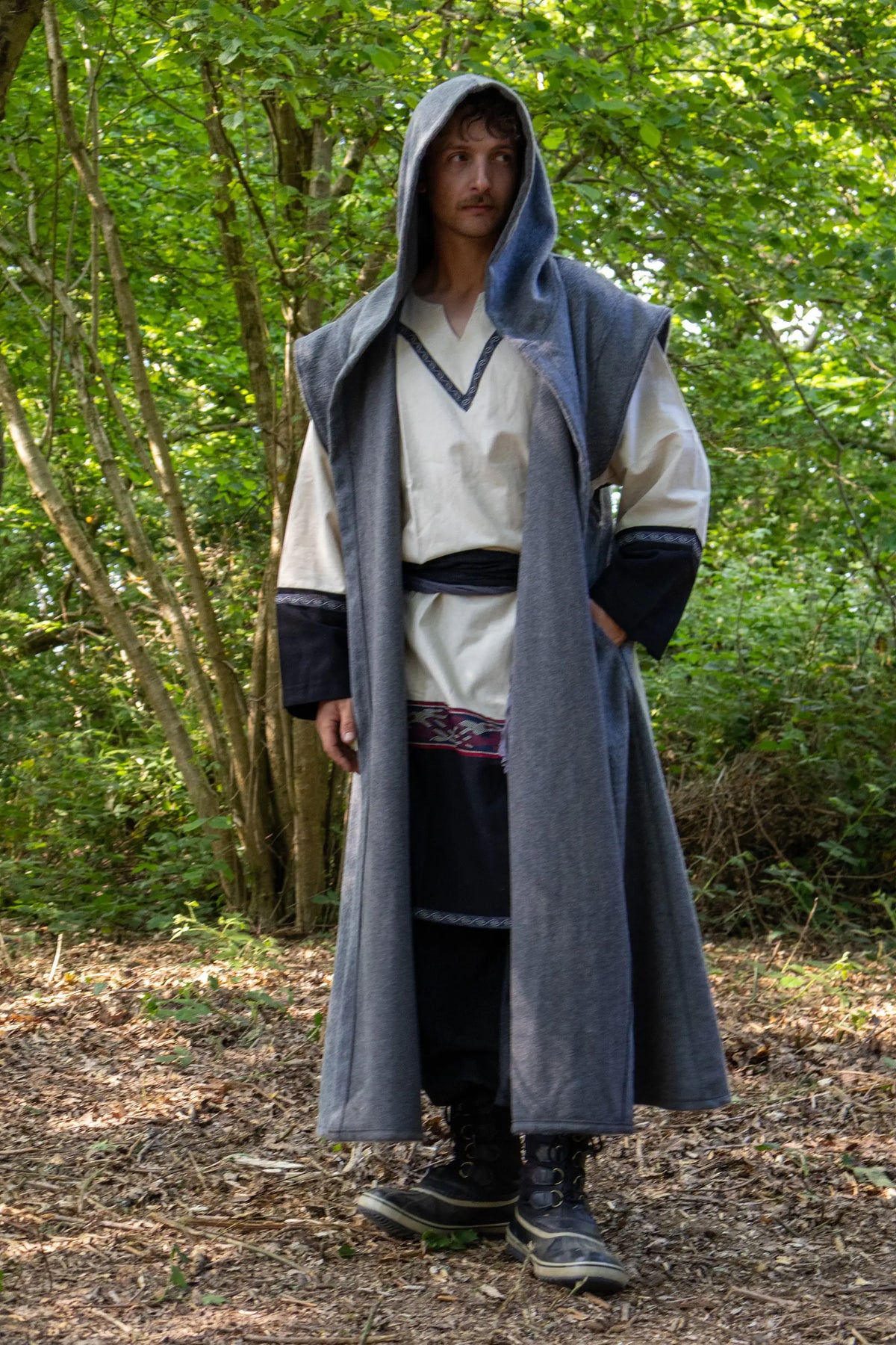 A man wearing a medieval-style long gray hooded waistcoat robe over a cream tunic and dark trousers, standing in a wooded outdoor setting.