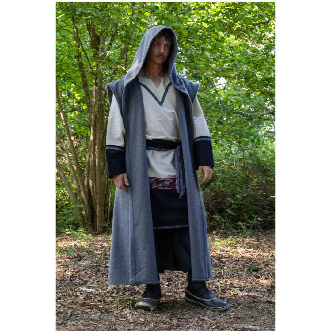 A man wearing a medieval-style long gray hooded robe over a white tunic and dark trousers, standing outdoors in a wooded area with trees and leaf-covered ground.