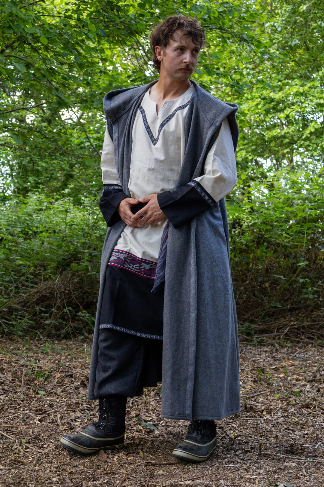 A man wearing a medieval-style costume with a long gray cloak, white tunic, and dark trousers, standing in a wooded outdoor setting.