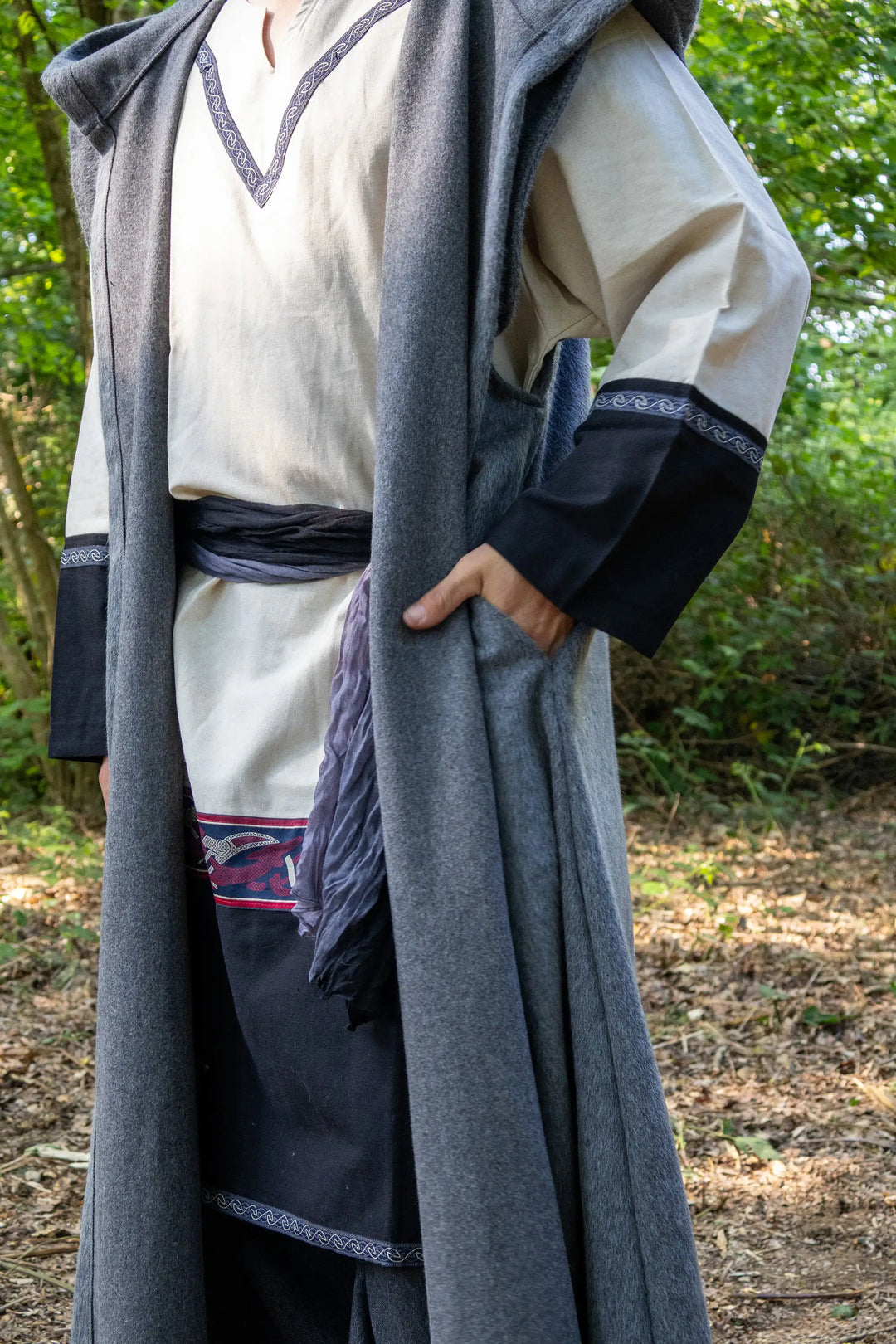 A person wearing a long medieval-style costume in an outdoor wooded setting. The outfit includes a cream tunic with dark trim, a dark blue apron, and a long gray cloak draped over the shoulders.