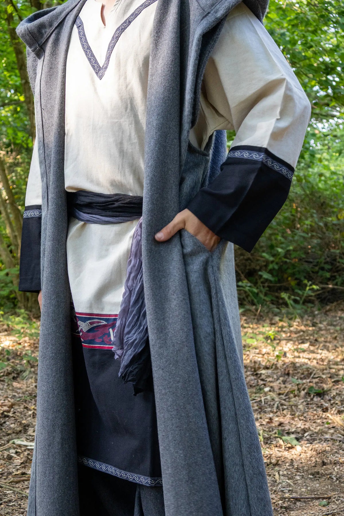 A person wearing a long medieval-style costume in an outdoor wooded setting. The outfit includes a cream tunic with dark trim, a dark blue apron, and a long gray cloak draped over the shoulders.