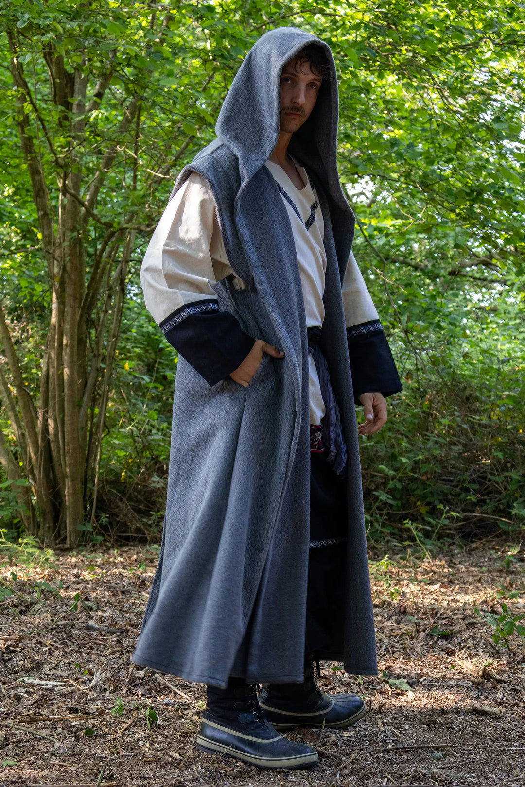 A person wearing a long gray hooded robe over a white and navy medieval-style tunic, standing outdoors in a wooded area with trees and leaf-covered ground.