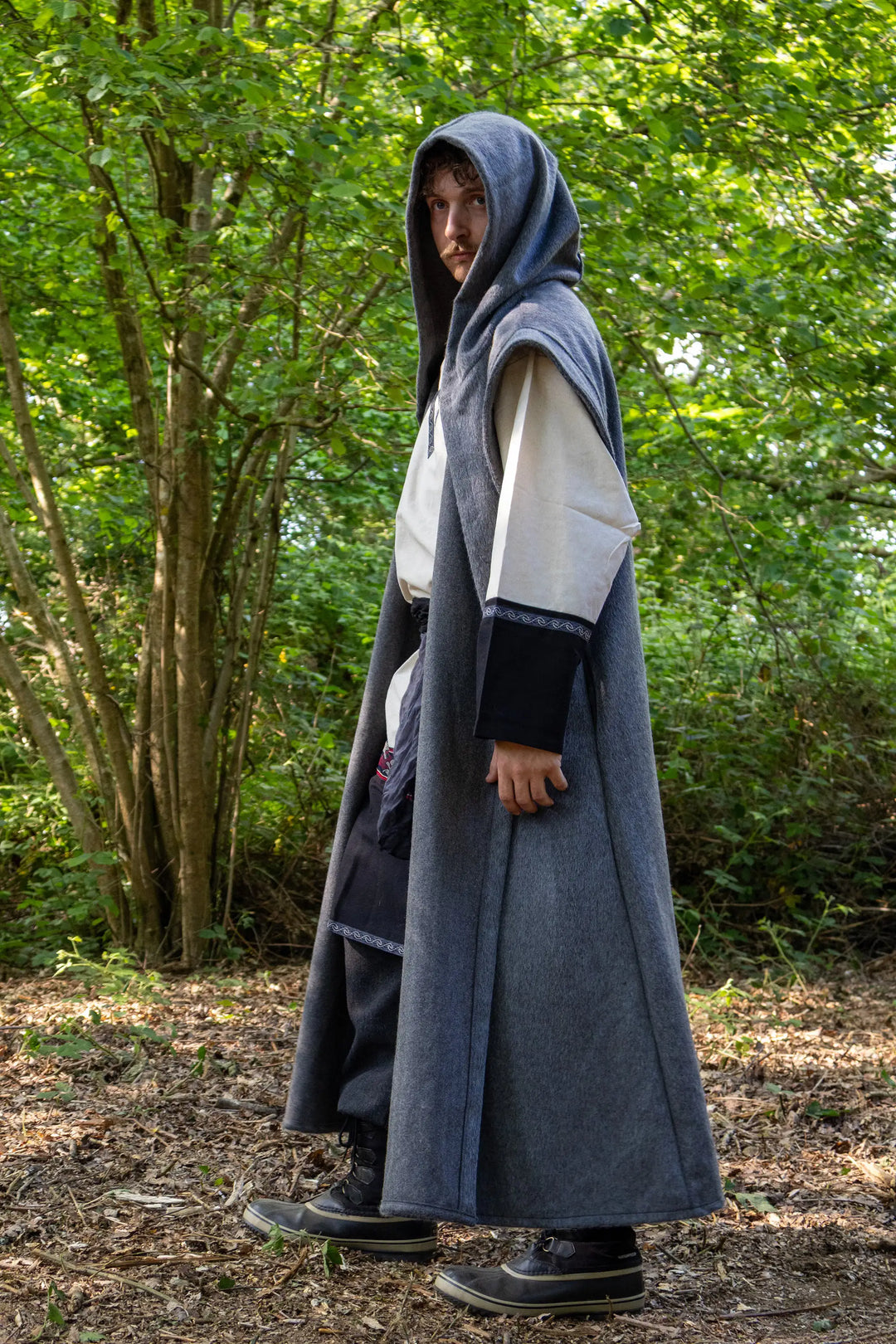 A person standing outdoors in a wooded area wearing a long grey hooded robe or cloak over a white tunic and dark pants.