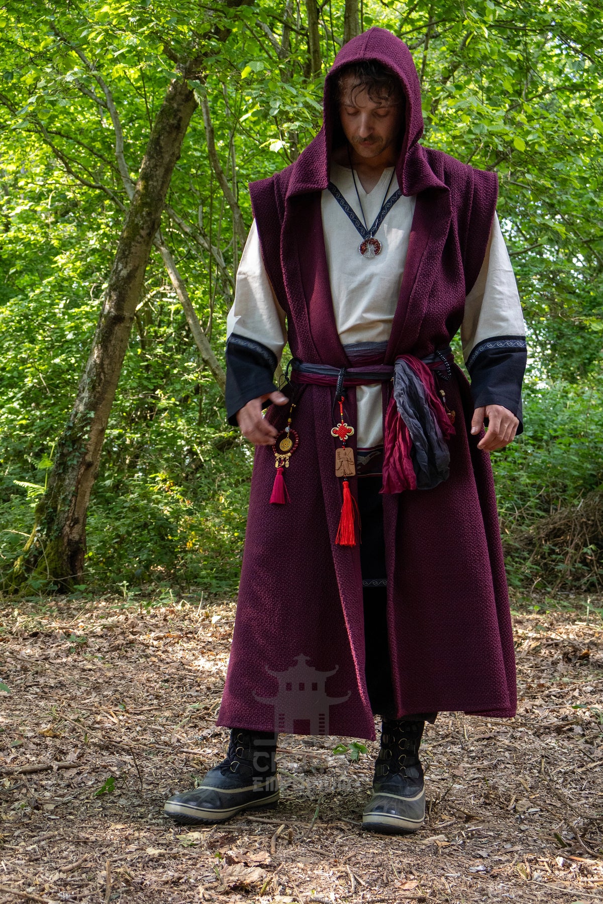 A man wearing a medieval-style costume stands in a wooded outdoor setting. The outfit features a long burgundy hooded cloak over a white tunic with dark black sleeves, accessorized with a dark sash, pouch, and tassel details.