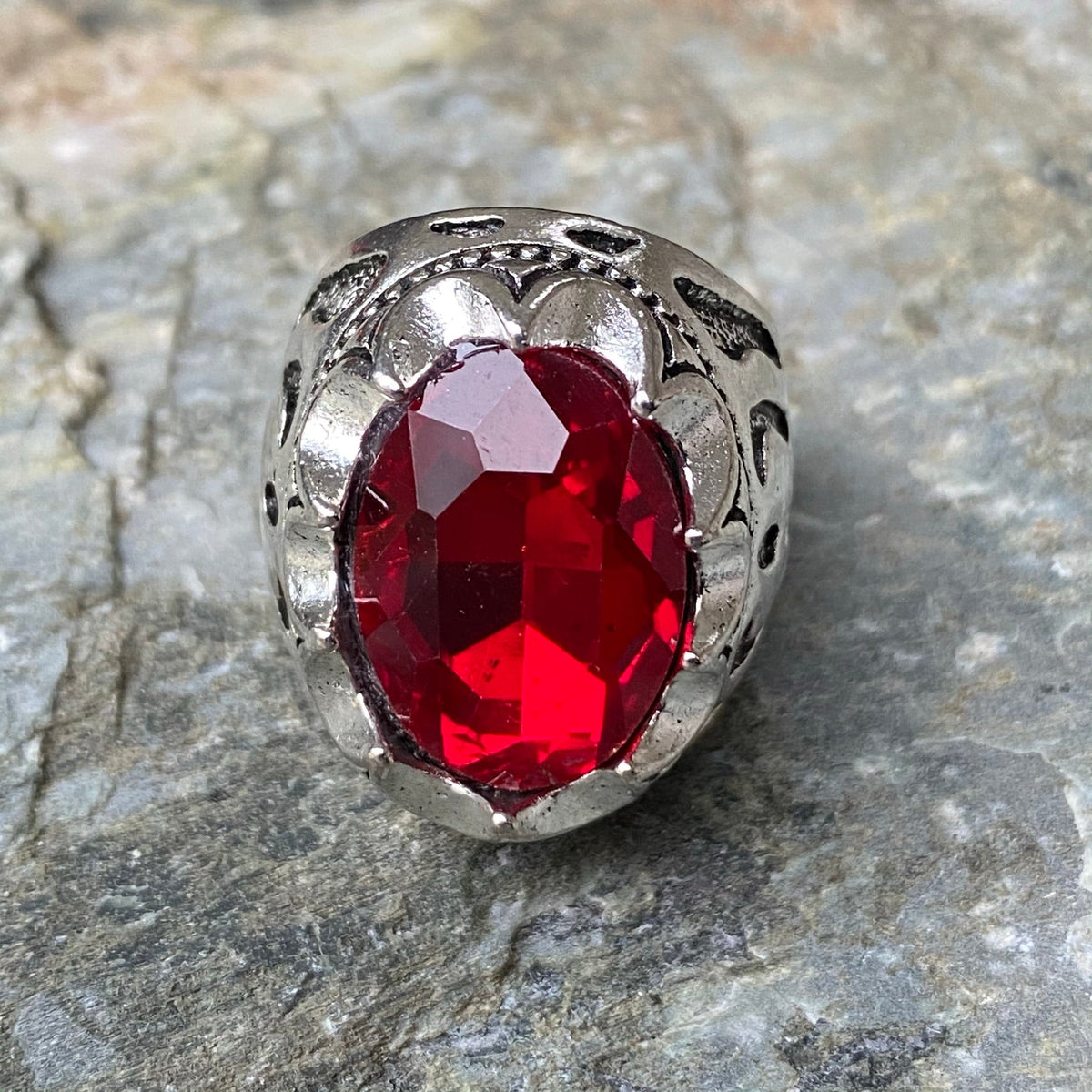 Silver LARP ring with ornate red gemstone, angled view on stone background.